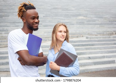 Education And People Concept. Portrait Of Two Smiling Multiethnic Students, Posing Outdoors. African American Male And European Female In The City, Study For Their Online Exams, Looking Away.