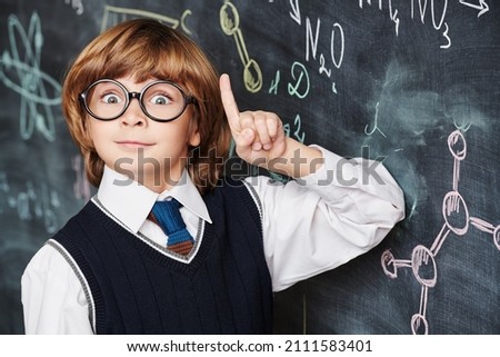 Education. Portrait of a cute smart boy in neat school uniform wearing glasses standing at the blackboard with chemical formulas and holding his index finger up. Clever kids.  