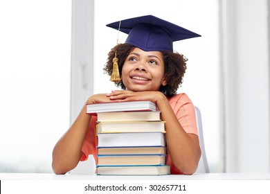 Education, School, Knowledge And People Concept - Happy Smiling African American Student Girl In Bachelor Cap With Books Sitting At Table And Dreaming At Home