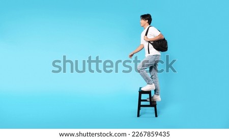 Education And Self Growth. Happy Korean Teen Guy Walking Up Stairs On Ladder Looking Aside Posing Holding Backpack Over Blue Studio Background. Panorama With Copy Space, Side View Shot