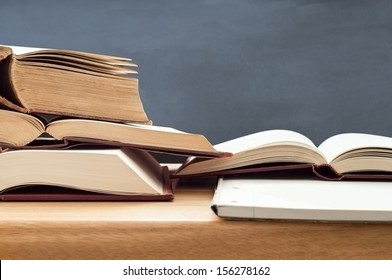 Education shot of stacked old books opened for study on wooden desk with blackboard in background.