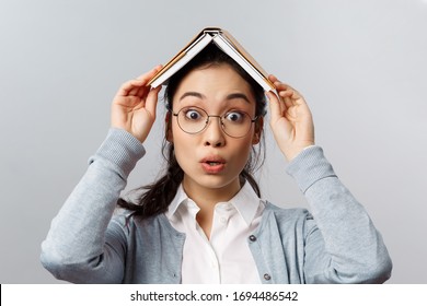 Education, University And People Concept. Close-up Portrait Of Wondred And Amused Asian Female In Glasses, Holding Planner, Notebook Or Book On Head, Realise Something Interesting, Grey Background