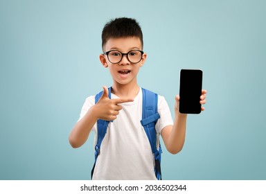 Educational App. Excited Asian Schoolboy Pointing At Smartphone With Blank Black Screen In His Hand, Male Kid With Backpack Wearing Eyeglasses Recommending New Application On Blue Background, Mockup