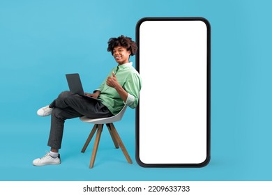 Educational App. Happy Black Student Guy Sitting With Laptop On Chair Near Big Blank Smartphone With White Screen And Showing Thumb Up, Male Teenager Recommending New Website Or Online Course, Mockup