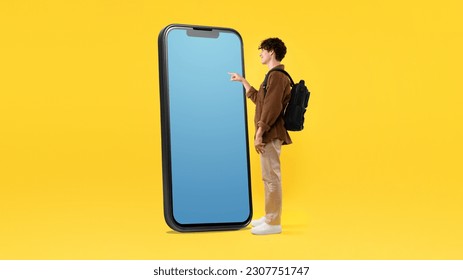 Educational Application. Student Guy With Backpack Using Giant Mobile Phone With Empty Screen, Touching Blank Gadget Touchscreen And Advertising New App Over Yellow Background. Panorama - Shutterstock ID 2307751747