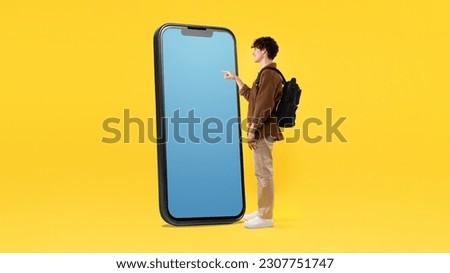 Educational Application. Student Guy With Backpack Using Giant Mobile Phone With Empty Screen, Touching Blank Gadget Touchscreen And Advertising New App Over Yellow Background. Panorama