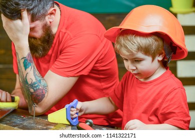 Educational Games Concept. Boy, Child Busy In Protective Helmet Learning To Use Handsaw With Dad. Father, Parent With Beard Looks Dissapointed While Son Play With Toy Saw.