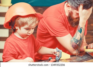 Educational Games Concept. Boy, Child Busy In Protective Helmet Learning To Use Handsaw With Dad. Father, Parent With Beard Looks Dissapointed While Son Play With Toy Saw.