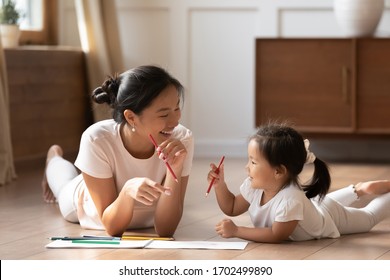 Educational pastime develop creativity skill in kid concept. Asian mother her small daughter lying on warm wooden floor in sunny cozy living room, mom teach girl paint use album and colourful pencils