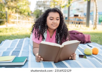 Educational Picnic. Relaxed Hispanic Student Woman Learning Reading Book Outdoors, Lying On Blanket In Green Campus Park Outside. Modern College Study And Homework Concept