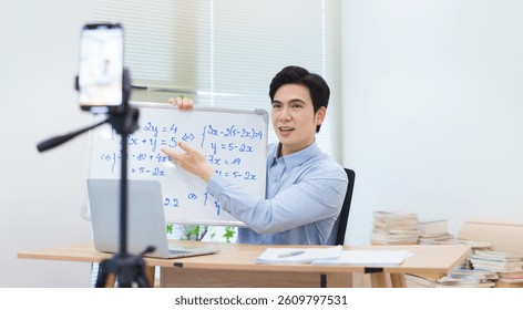 Educator enthusiastically demonstrates math problems on a whiteboard while conducting an online lesson from a well-organized office space with natural light.