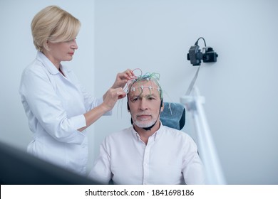 EEG Examination. Female Neurologist Adjusting Electrodes On Gray-haired Male Patient Head Undergoing Electroencephalogram