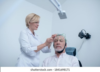 EEG Examination. Female Neurologist Placing Electrode Cap On Gray-haired Male Patient Head