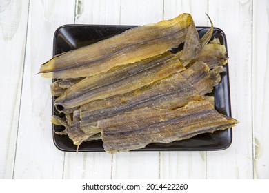 Eel (Latin Anguilla) Dried Cut Into Strips On A Black Rectangular Plate With A Wooden Table Background. Food Is A Delicacy Snack For Beer.