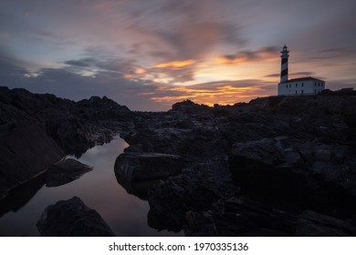 An Eerie Scenery Of A Lighthouse Under The Pink And Golden Sky In The Rocky Bay Of The Ocean