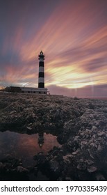 An Eerie Scenery Of A Lighthouse Under The Pink And Golden Sky In The Bay Of The Ocean