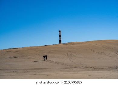 An Eerie View Of A Sandy Desert With Two Visitors Heading To The Lonely Lighthouse