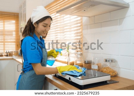 Efficient Asian housewife employs a rag and sprayer to meticulously clean the electric stove in her kitchen, showcasing her dedication to maintaining a sparkling and well-maintained cooking space.