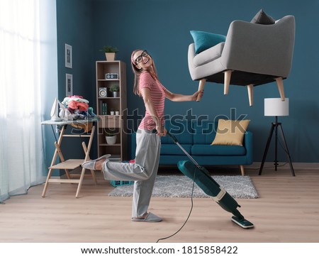 Efficient happy housewife cleaning up her apartment: she is lifting the armchair with one hand and vacuuming the floor, effortless easy housekeeping concept