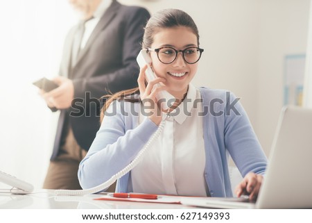 Efficient smiling secretary answering phone calls and talking with customers, she is sitting at desk and working with a laptop