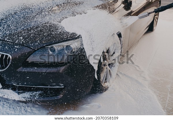 Efficient vehicle cleaning, spraying detergent\
on vehicle with high-pressure washer at car wash center. Outdoor\
car cleaning concept. Black surface in suds during manual exterior\
carwashing