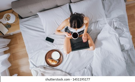 Efficient Young Millennial Girl Sitting on a Bed in the Morning, Uses Laptop Computer and Eats Croissants and Drinks Coffee for Breakfast. Top Down Shot. - Powered by Shutterstock