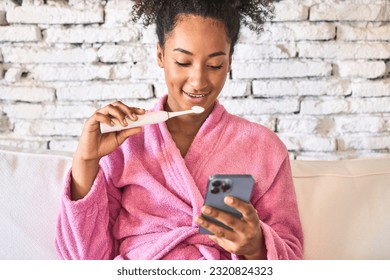 Efficient young woman multitasks: brushing teeth, messaging friend in cozy robe on sofa. - Powered by Shutterstock