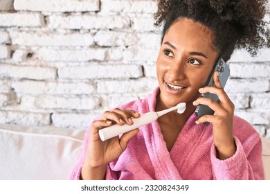 Efficient young woman multitasks: brushing teeth, messaging friend in cozy robe on sofa. - Powered by Shutterstock