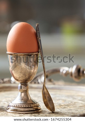 Egg in a silver eggcup and a spoon on wooden table. Vertical portrait of the set and a fresh full protein brown egg. Blur background, space.