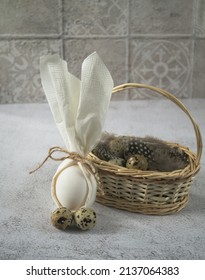 An Egg Is Wrapped In A Paper Napkin And Tied With A Natural Ribbon In The Shape Of A Bunny Next To A Natural Wood Basket With Quail Eggs On A Gray Background.