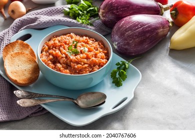 Eggplant caviar in blue bowl and fresh vegetables on background.