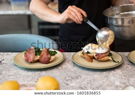 Eggs benedict with crisp bacon and spinach being topped with hollandaise sauce for morning brunch.