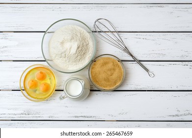 Eggs, Flour, Milk, Brown Sugar With Whisk On White Wooden Table From Above. 