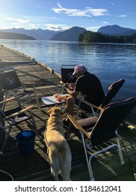 Egmont, British Columbia, Canada - July 2nd, 2018: A Man Eating Dinner On The Dock With His Yellow Lab In Gorgeous Egmont, B.C., Waiting On The Rest Of The Family To Join