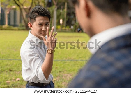 An egotistical and cocky young asian man taunts his workmate with a mocking hand gesture. Rivals or enemies in the workplace.