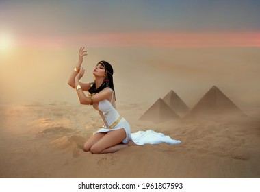 Egypt Style Woman. Sexy beautiful girl goddess Queen Cleopatra sits on sand desert pyramids. Art ancient pharaoh costume white dress gold accessories. Egyptian makeup. hands raised to sky lady praying