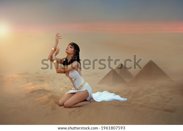 Egypt Style Woman. Sexy beautiful girl goddess\
Queen Cleopatra sits on sand desert pyramids. Art ancient pharaoh\
costume white dress gold accessories. Egyptian makeup. hands raised\
to sky lady praying