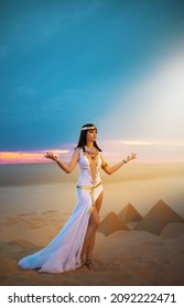 Egypt Woman pagan prays hands raised to heaven blue sky. Sexy girl Egyptian goddess Queen Cleopatra. yellow sand Sahara desert pyramids. Art ancient pharaoh costume white dress gold accessories crown