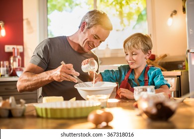 A Eight Years Old Blond Boy Is Cooking  With His Father In A Luminous Kitchen. They Are Sitting At A Wooden Table The Dad Is Mixing The Preparation While His Son Is Adding Ingredients.Shot With Flare