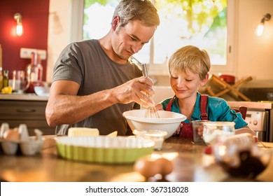 A Eight Years Old Blond Boy Is Cooking  With His Father In A Luminous Kitchen. They Are Sitting At A Wooden Table The Dad Is Mixing The Preparation While His Son Is Adding Ingredients.Shot With Flare