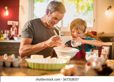 A Eight Years Old Blond Boy Is Cooking  With His Father In A Luminous Kitchen. They Are Sitting At A Wooden Table The Dad Is Mixing The Preparation While His Son Is Adding Ingredients.Shot With Flare