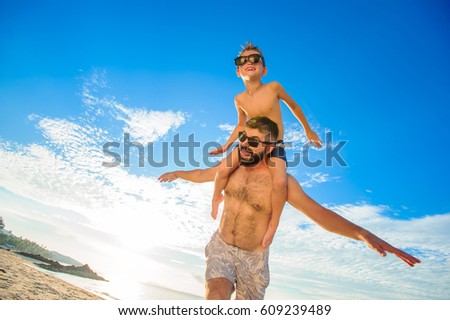 Eight years old boy sitting on dad's shoulders. Both in swimming shorts and sunglasses, having fun on the beach. Bottom view. Blue sky and altocumulus clouds behind them