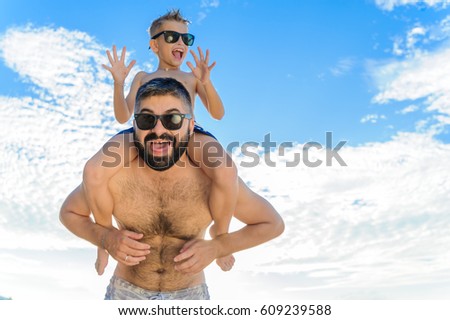 Eight years old boy sitting on dad's shoulders. Both in swimming shorts and sunglasses, having fun on the beach. Bottom view. Blue sky and altocumulus clouds behind them