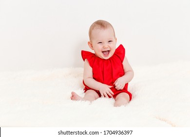 an eight-month-old cute Caucasian baby girl in a red suit sits on the bed and laughs. children under one year old.