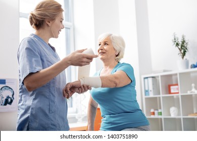 Elastic bandage. Pleasant delighted nurse looking at her patient while holding a white bandage - Powered by Shutterstock