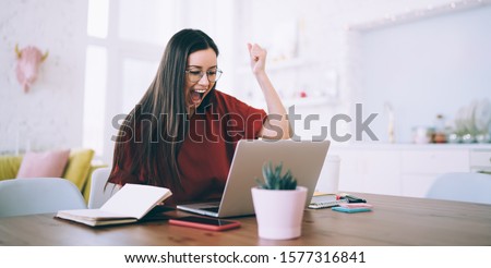 Elated excited successful female with long hair raising fist and screaming for celebrating victory watching on laptop at cozy apartment