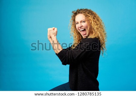 Elated satisfied young redhead curly woman celebrating and cheering a success while punching the air with her fists over blue wall background. Yes. I did it. Positive feelings. Studio shot.