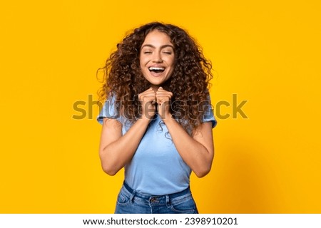Elated woman with curly hair, eyes closed, fists clenched in a gesture of victory or celebration, laughing with joy against a yellow background