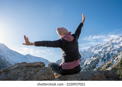 Elated woman with hands outstretched like wings on a vantage in snowy mountains on a sunny day. She is sitting on a boulder. - Shutterstock ID 2220937021