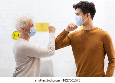 Elbow Bump Greeting. Asian Guy And Senior Woman In Medical Masks Bumping Elbows Meeting In Hospital Or Office Indoors. Social Distancing, Avoiding Handshake And Coronavirus Infection Spread Concept